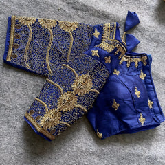 Traditional royal blue silk blouse featuring intricate gold embroidery and beadwork with floral and paisley motifs on the sleeves and neckline, displayed flat on a textured surface with a bunch of artificial flowers and a woven mat in the background.