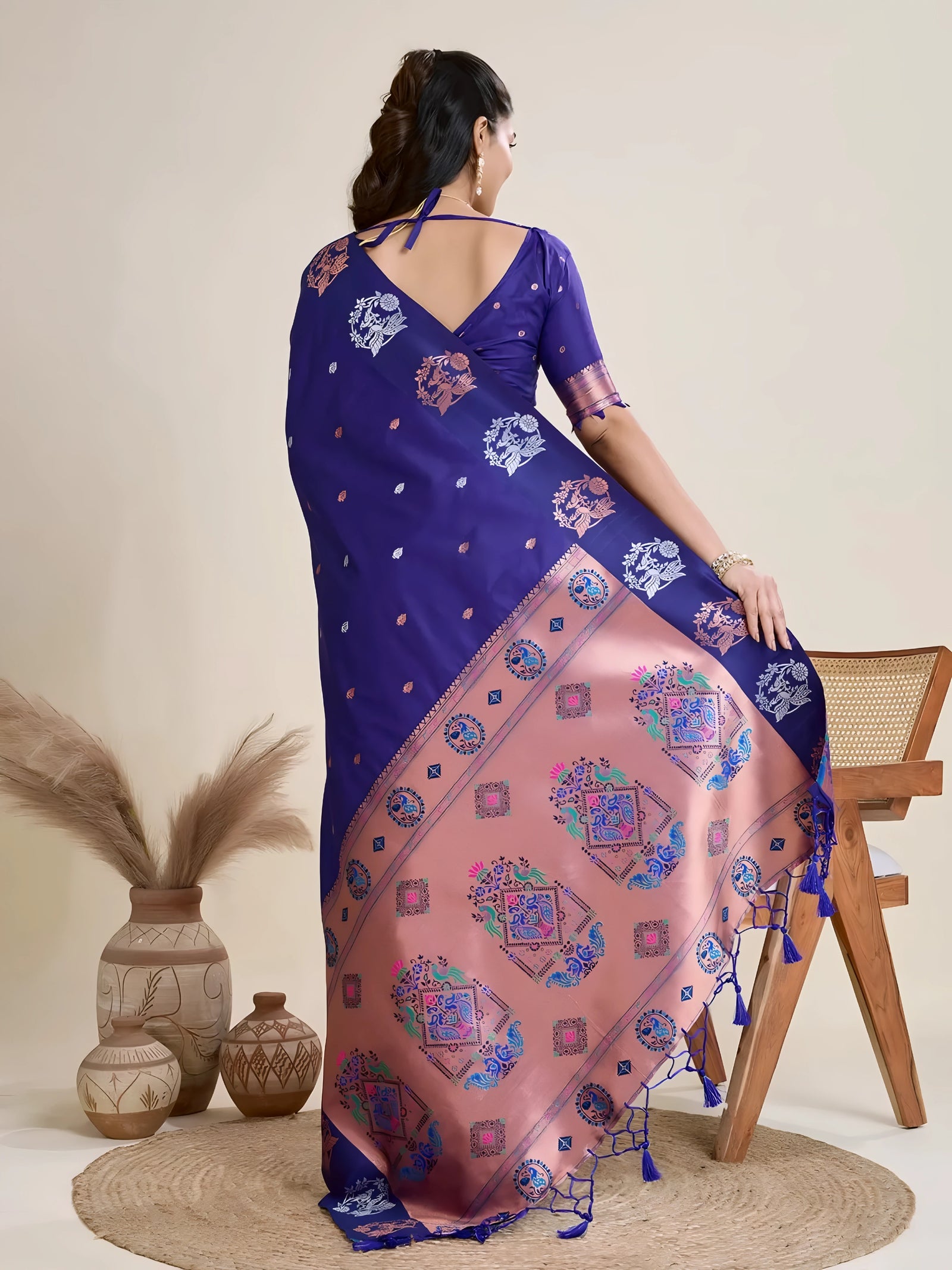 A woman models a luxurious Royal Blue Soft Silk Saree. The saree features silver Zari motifs on the body and a wide pallu in contrasting rose gold/peach, richly woven with multi-colored peacock and floral designs. She wears a matching short-sleeve blue blouse and heavy gold Kundan jewelry, posing indoors.