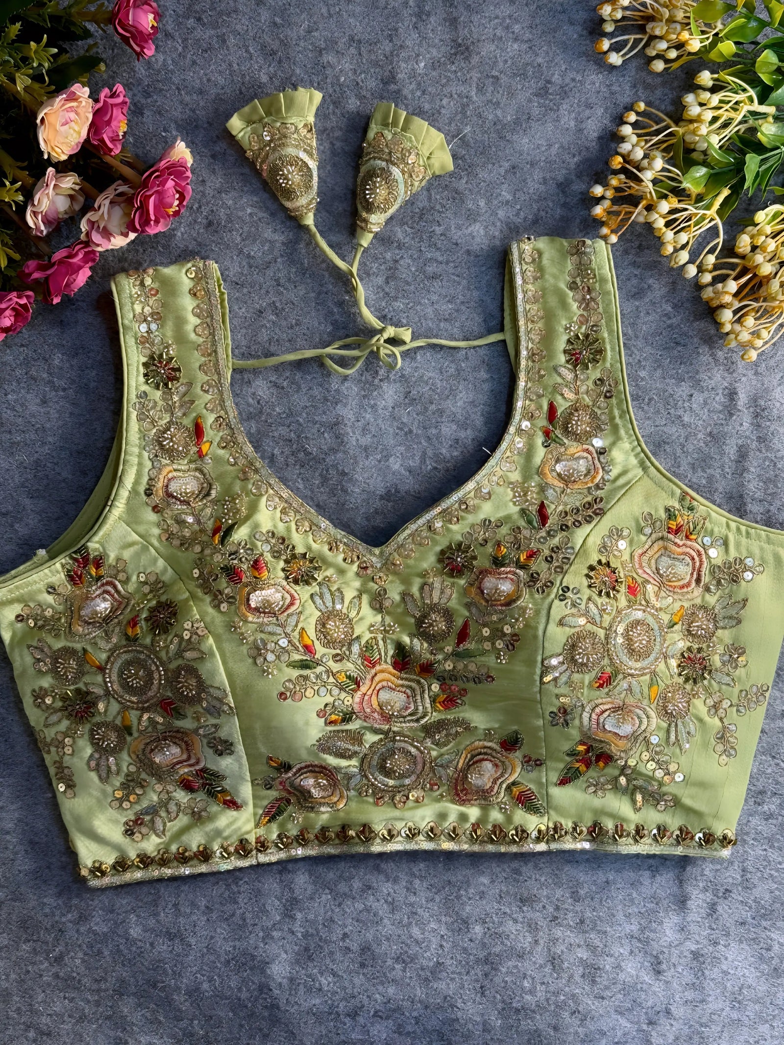 Traditional sage green silk blouse featuring intricate gold zardozi embroidery and beadwork with multicolor floral and leaf motifs on the front and neckline, finished with decorative tassel ties, displayed flat on a textured grey background with artificial pink and yellow flowers in the corners.