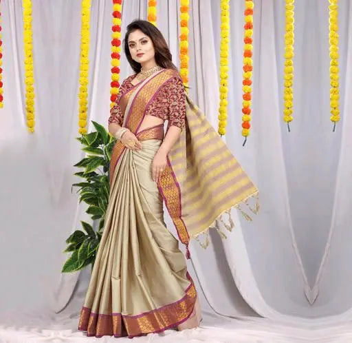 Woman wearing a light beige silk saree with maroon blouse and yellow striped pallu, standing in front of decorative yellow and orange floral garlands and green plants
