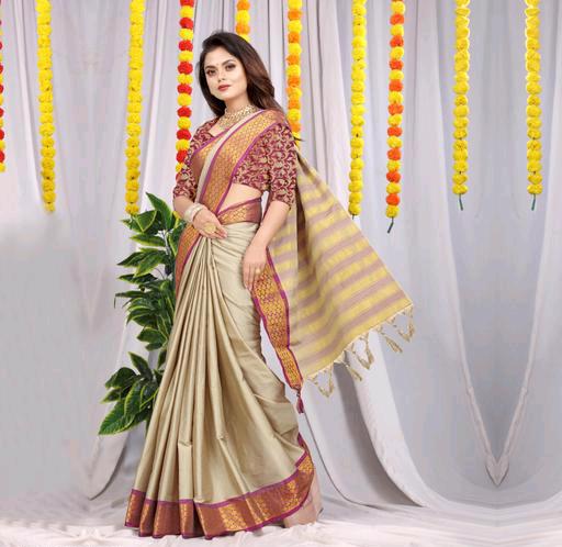 Woman wearing a light beige silk saree with maroon blouse and yellow striped pallu, standing in front of decorative yellow and orange floral garlands and green plants