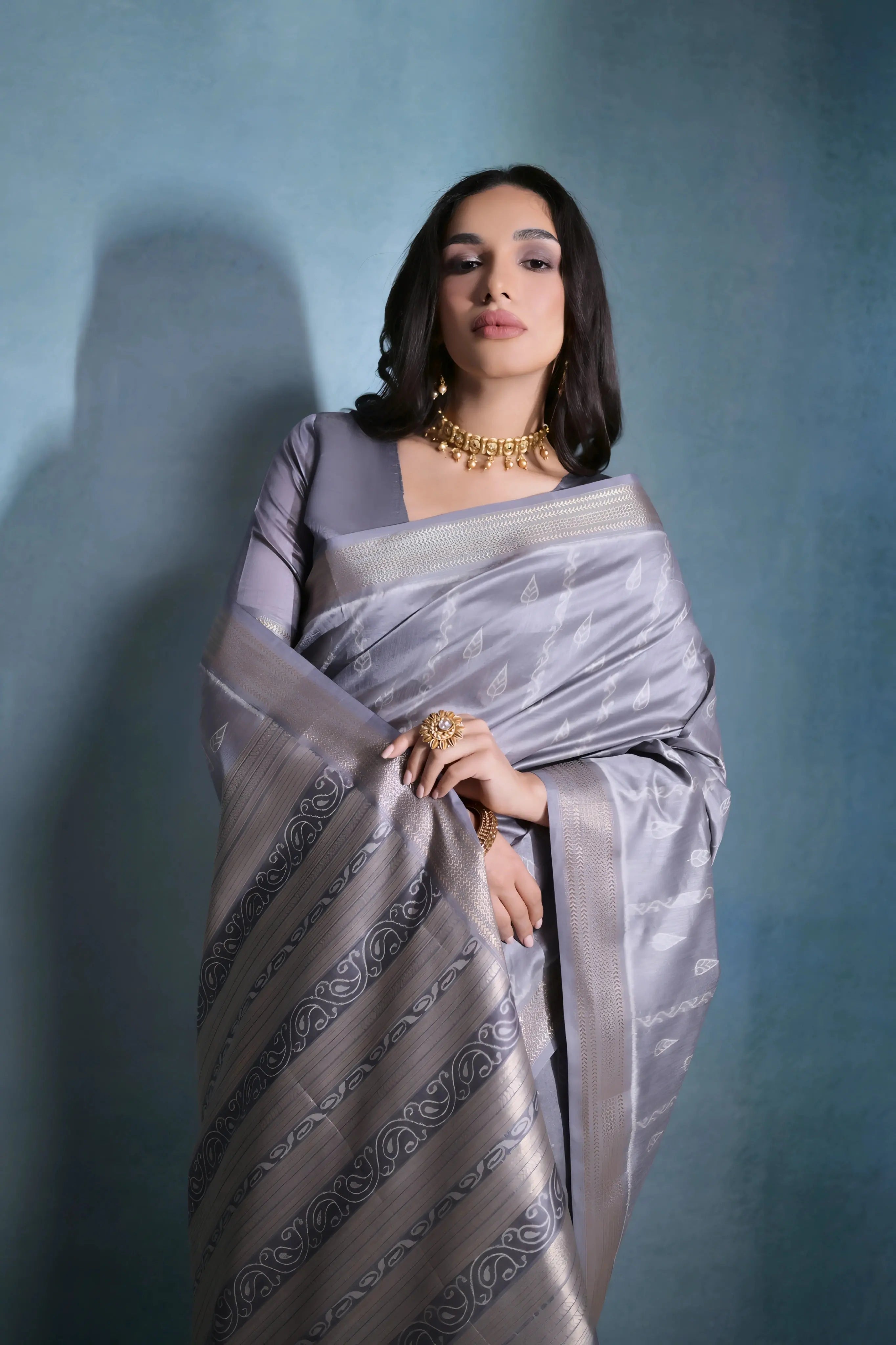 A woman models an elegant light Grey Soft Raw Silk Saree. The saree body has subtle woven motifs, and the wide pallu features intricate horizontal bands of metallic Zari weaving and traditional Ikat-style geometric patterns in light and dark grey. She wears a matching short-sleeve grey blouse and gold Kundan jewelry, posing indoors.
