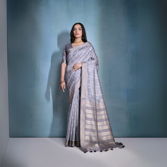 A woman models an elegant light Grey Soft Raw Silk Saree. The saree body has subtle woven motifs, and the wide pallu features intricate horizontal bands of metallic Zari weaving and traditional Ikat-style geometric patterns in light and dark grey. She wears a matching short-sleeve grey blouse and gold Kundan jewelry, posing indoors.