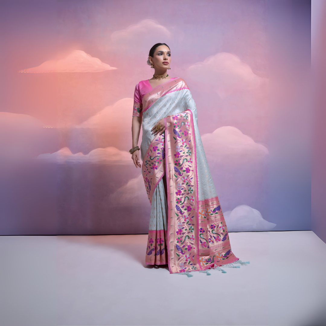 A woman models an elegant Light Blue/Icy Gray Soft Banarasi Silk Saree. The saree features a wide, vibrant Fuchsia Pink border and pallu with dense, colorful floral Kanjivaram-style weaving. She wears a contrasting short-sleeve pink blouse and a gold choker necklace, posing indoors against a surreal pink and blue cloud backdrop.