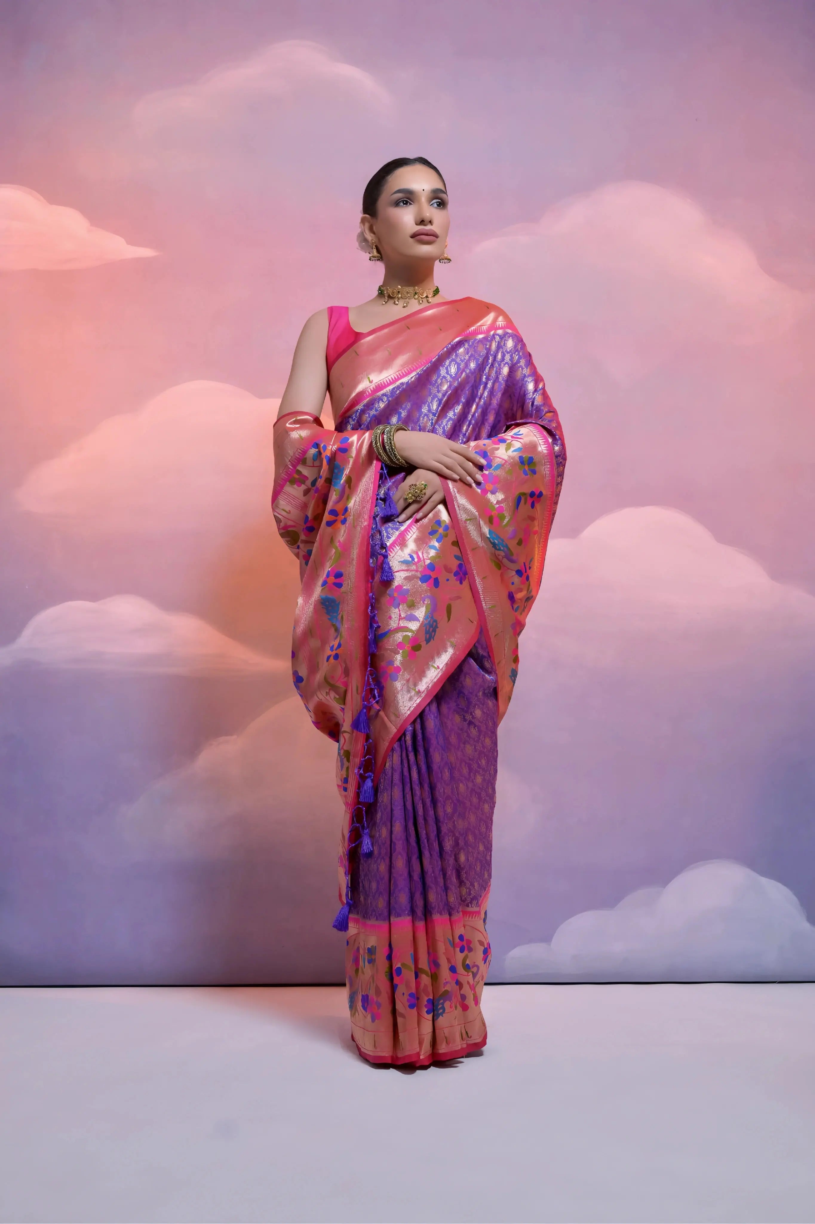 A woman models a magnificent Dual-Toned Purple and Rose Gold Soft Banarasi Silk Saree. The saree features a wide, vibrant Fuchsia Pink border and pallu with dense, colorful peacock and floral Kanjivaram-style weaving. She wears a contrasting pink sleeveless blouse and a heavy gold Kundan choker necklace.
