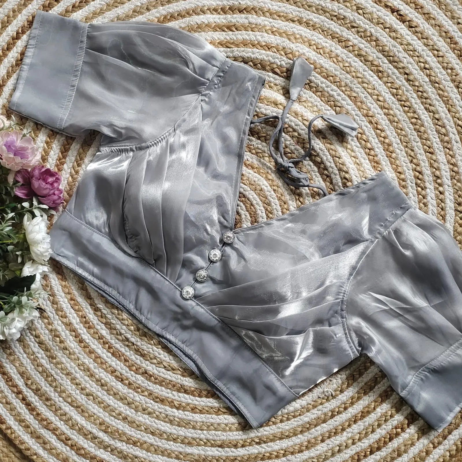 Silver satin blouse with pleated detailing and four decorative silver buttons on the front, featuring elbow-length sleeves and tie-up tassels, displayed on a circular woven mat with white and pink flowers on the side.
