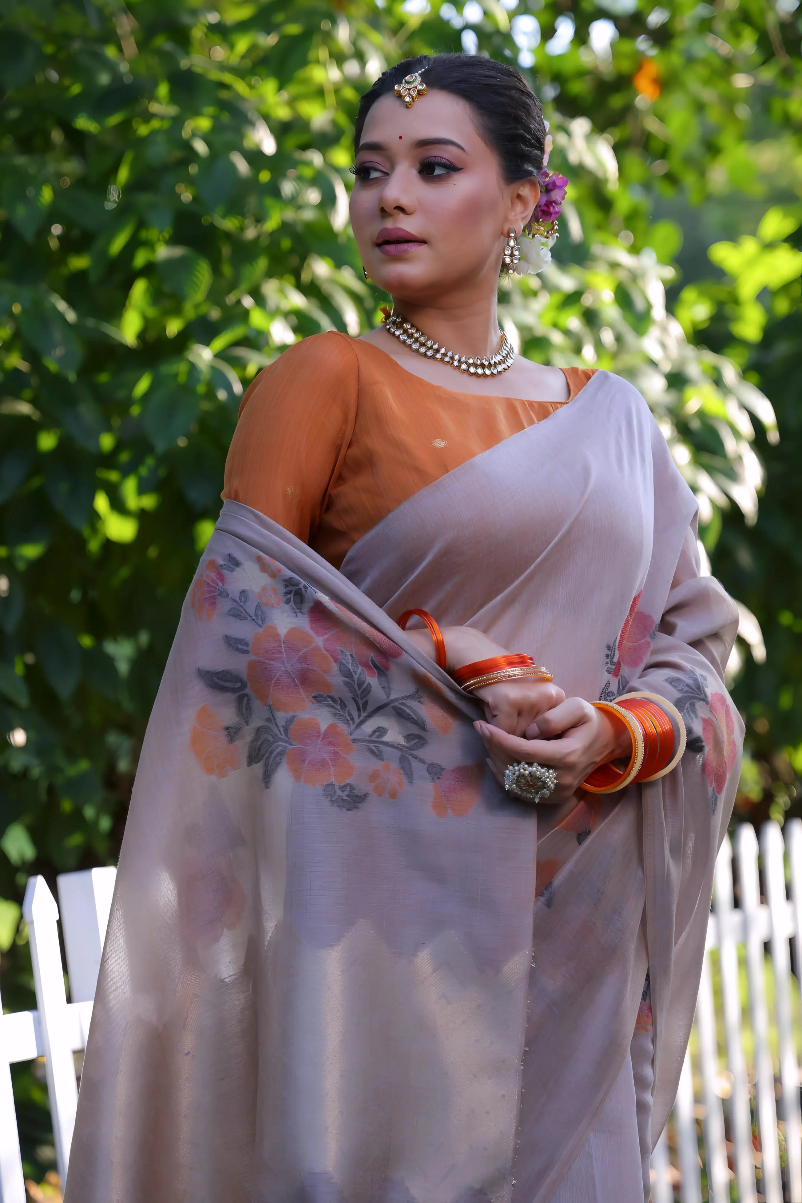 A woman models an elegant Dusty Gray Soft Muga Cotton Saree. The pallu features large, artistic Resham floral weaving in pink and coral tones. She wears a contrasting rust orange blouse and is accessorized with a pearl choker necklace and vibrant orange bangles, posing outdoors by a white fence.