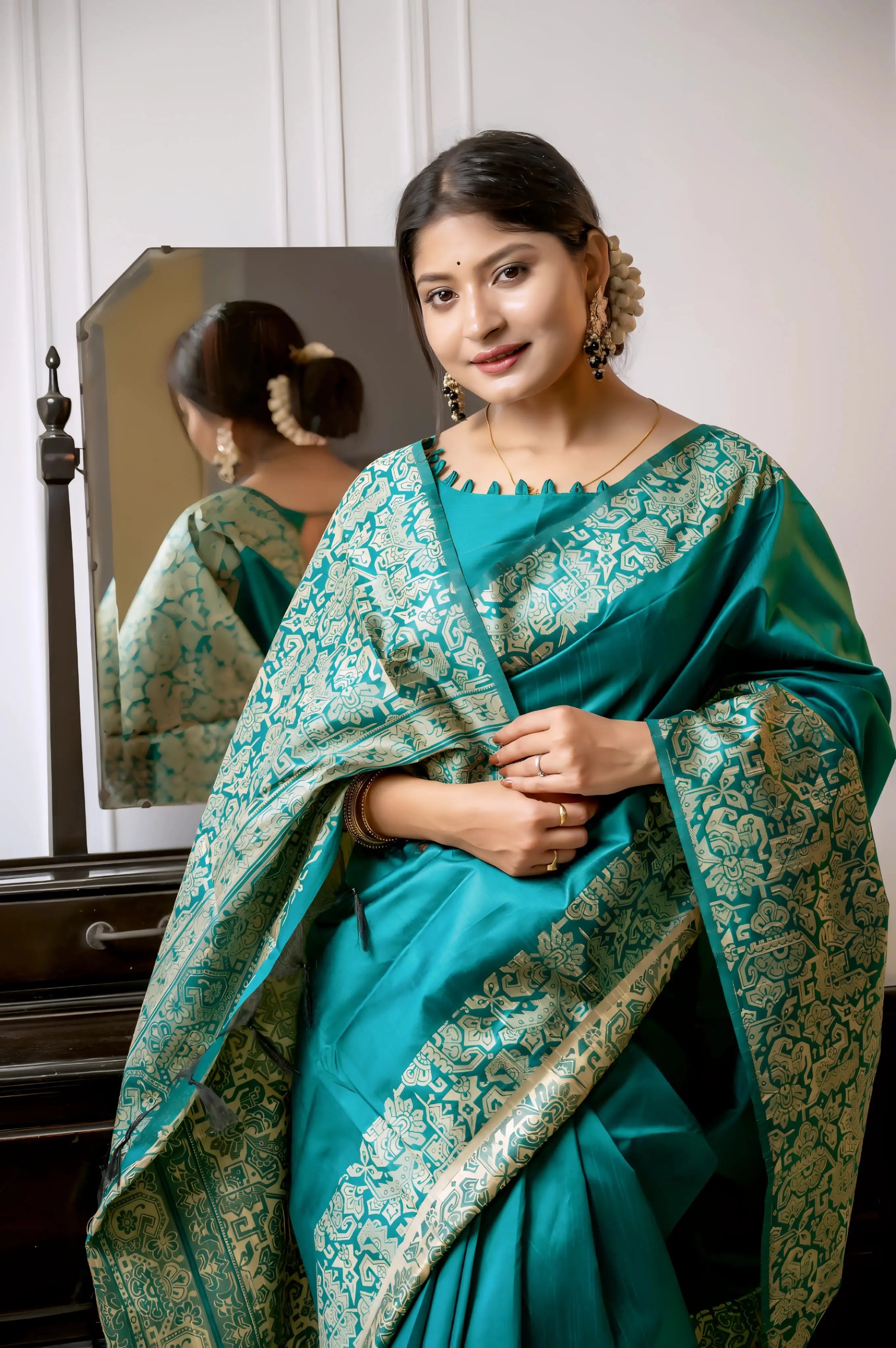 A woman models a luxurious Deep Sea Green Raw Silk Saree. The saree features a wide border and pallu with dense, intricate gold Zari weaving in a Kalamkari-style floral and paisley pattern. She wears a matching short-sleeve blouse with a scalloped neckline and heavy traditional gold jewelry, posing indoors next to a wooden vanity.