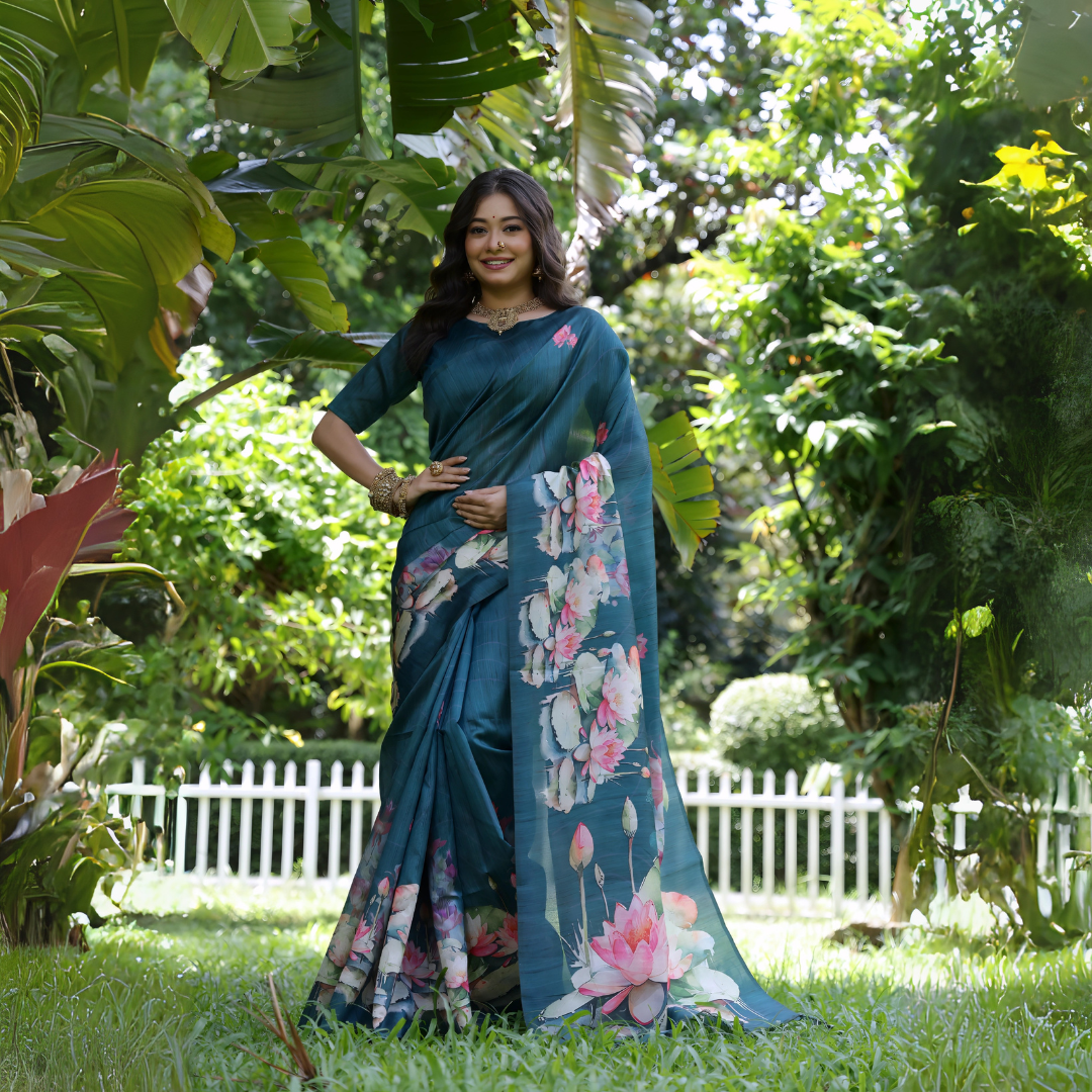 A woman models a stunning Deep Rama Blue Soft Tussar Silk Saree. The saree features a large-scale digital print of colorful floral and lotus motifs in pink, white, and green on the pallu and lower drape. She wears a matching elbow-sleeve blue blouse and heavy gold Kundan jewelry, posing outdoors in a garden setting.