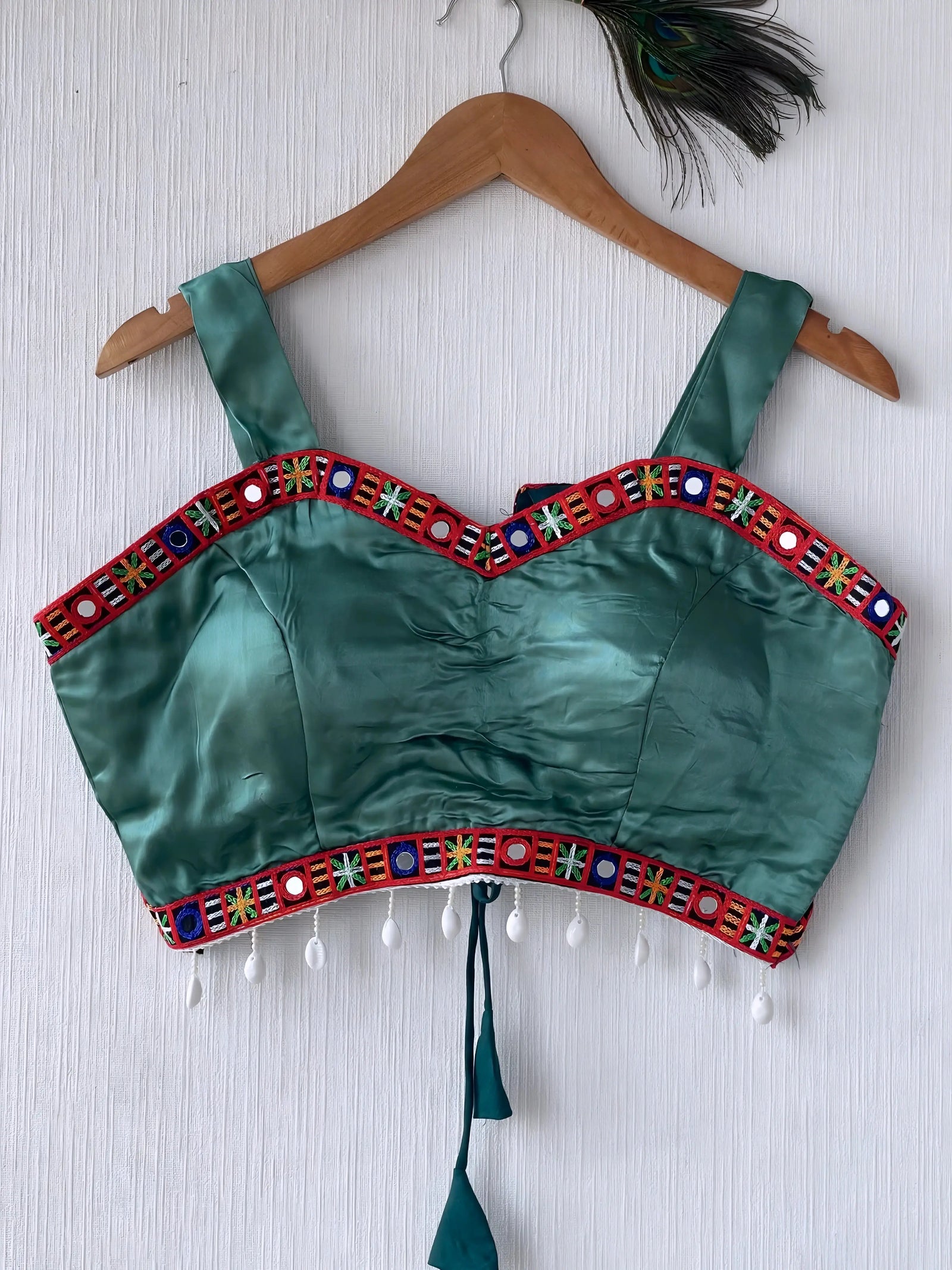 Teal green ethnic blouse featuring vibrant mirror work and a multicolored embroidered geometric border in red, green, blue, and white, adorned with white pearl drop tassels and a tie-up waist detail. Displayed on a wooden hanger against a textured off-white wall with peacock feather, creating a festive and traditional look.