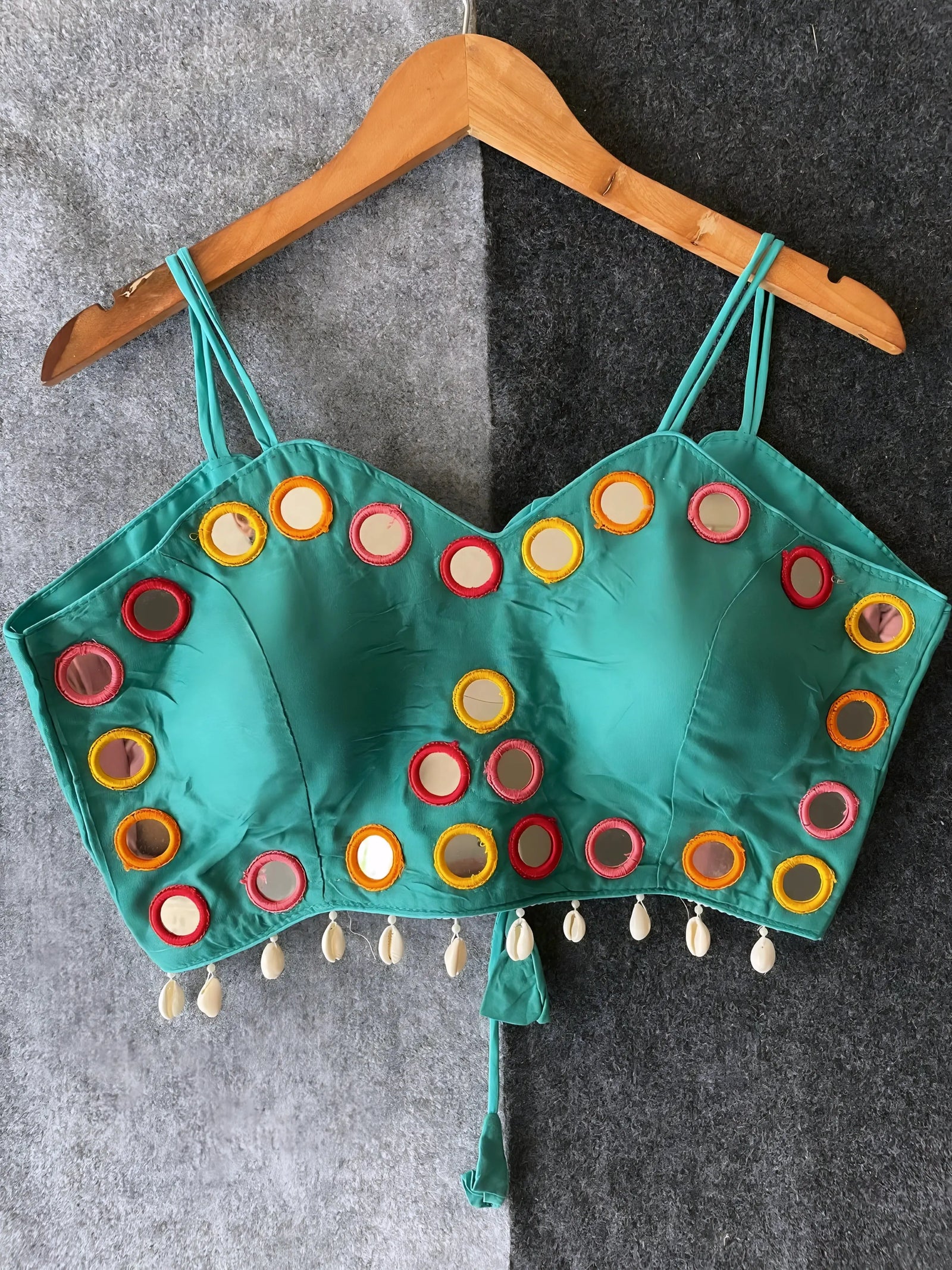 Teal corset-style blouse embellished with vibrant round mirrors and multicolored embroidered borders, featuring thin shoulder straps and shell tassel drops along the hem. Displayed on a wooden hanger against a two-tone grey textured background, showcasing its festive and modern ethnic