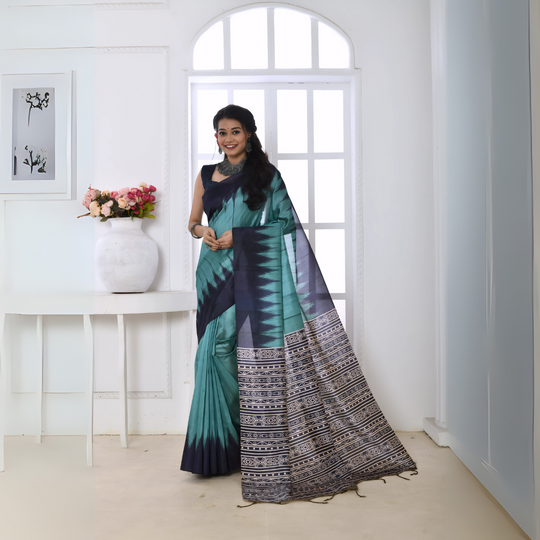 A woman models a vibrant Sky Blue/Teal Soft Tussar Silk Saree. The saree features a bold black/navy zigzag pattern along the border and transitions on the body. The pallu has a wide panel of dense tribal or Ikat-style geometric print in light grey, black, and white. She wears a dark sleeveless blouse and chunky oxidized silver jewelry, posing indoors by a large arched window.