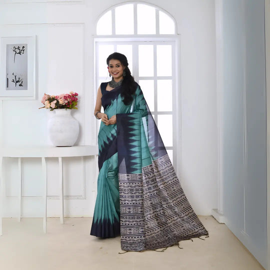 A woman models a vibrant Sky Blue/Teal Soft Tussar Silk Saree. The saree features a bold black/navy zigzag pattern along the border and transitions on the body. The pallu has a wide panel of dense tribal or Ikat-style geometric print in light grey, black, and white. She wears a dark sleeveless blouse and chunky oxidized silver jewelry, posing indoors by a large arched window.