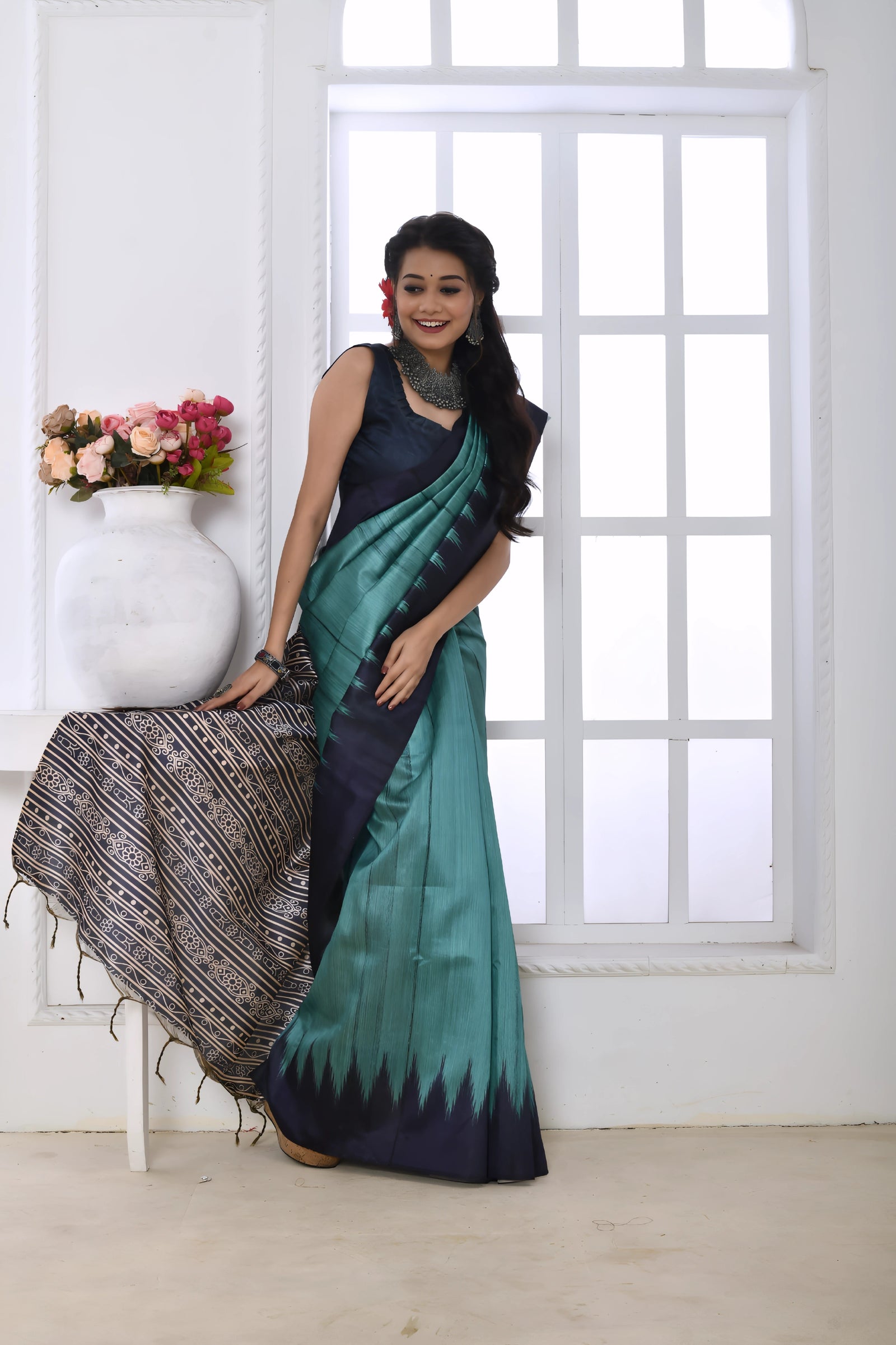 A woman models a vibrant Sky Blue/Teal Soft Tussar Silk Saree. The saree features a bold black/navy zigzag pattern along the border and transitions on the body. The pallu has a wide panel of dense tribal or Ikat-style geometric print in light grey, black, and white. She wears a dark sleeveless blouse and chunky oxidized silver jewelry, posing indoors by a large arched window.