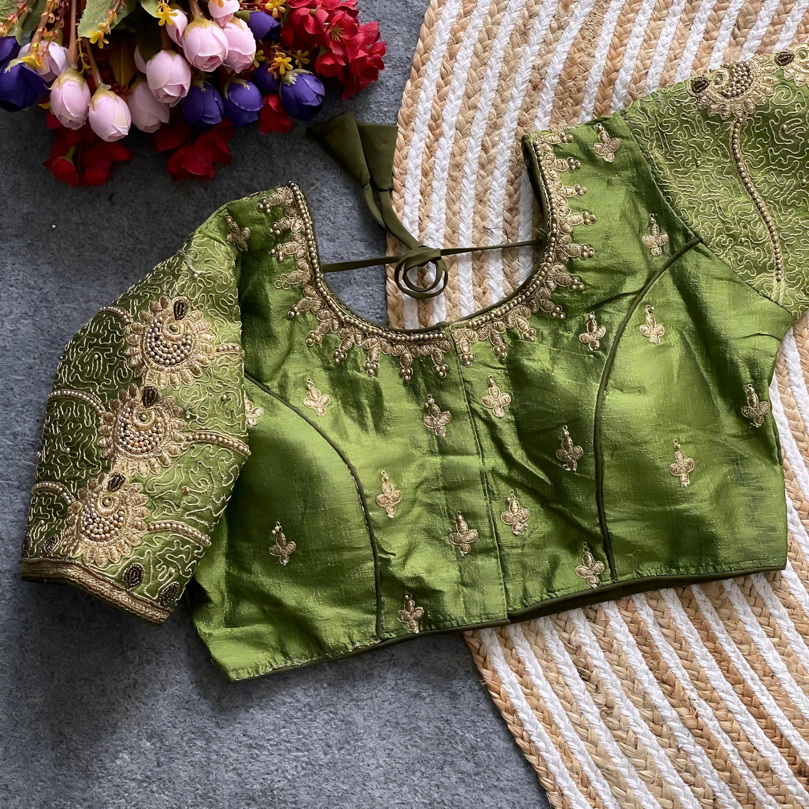 Traditional teal silk blouse featuring intricate gold embroidery and beadwork with floral and paisley motifs on the sleeves and neckline, displayed flat on a textured surface with a bunch of artificial flowers and a woven mat in the background.