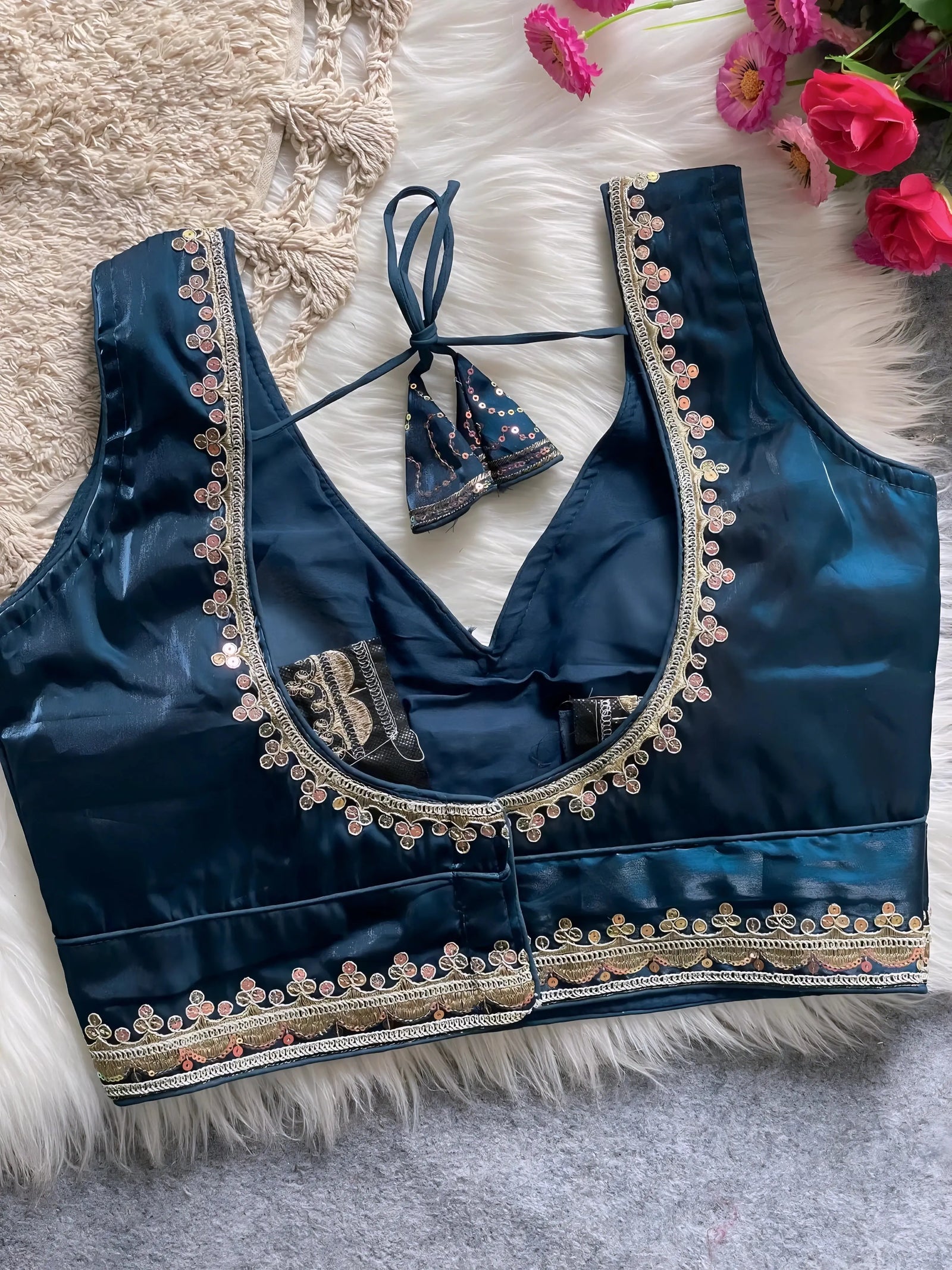 Traditional teal silk blouse featuring intricate gold sequin and thread embroidery along the neckline and hem, with decorative tassel tie at the back, displayed flat on a soft fur and textured mat background with artificial pink flowers in the corner.