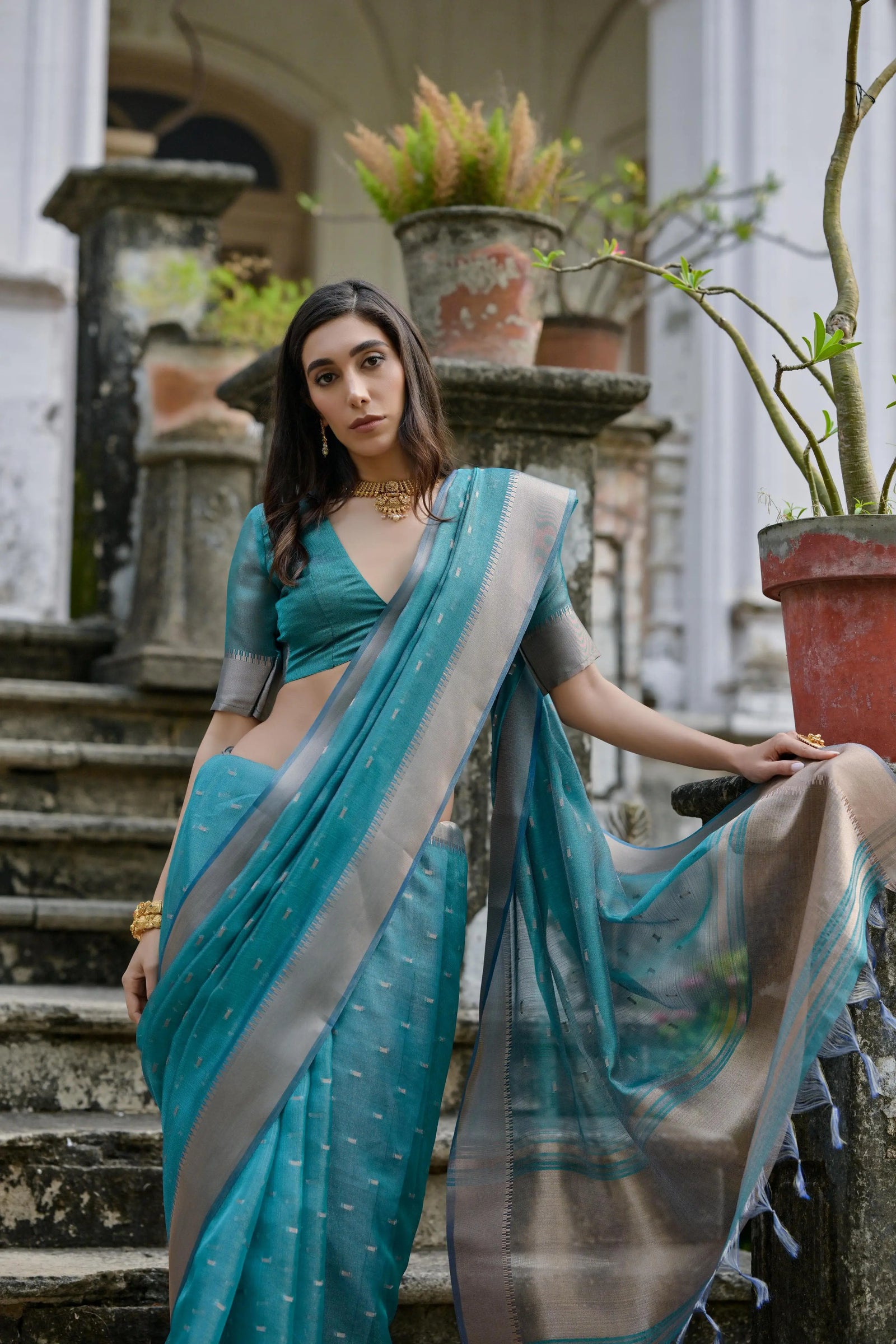 A woman models a stunning Deep Sea Blue Soft Silk Saree. The saree features scattered metallic motifs and a wide, contrasting silver/gray Zari woven border. She wears a matching blue elbow-sleeve blouse and a delicate gold choker necklace, posing on an ornate stone staircase.