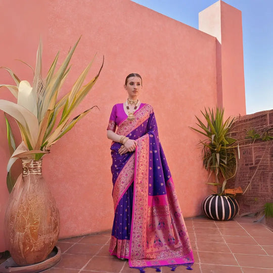 A woman models a vibrant Banarasi Silk Saree. The saree is royal purple with gold Zari weaving across the body, featuring a wide, ornate hot pink and gold Zari woven border and pallu. She pairs it with a short-sleeve pink blouse and heavy traditional gold jewelry.