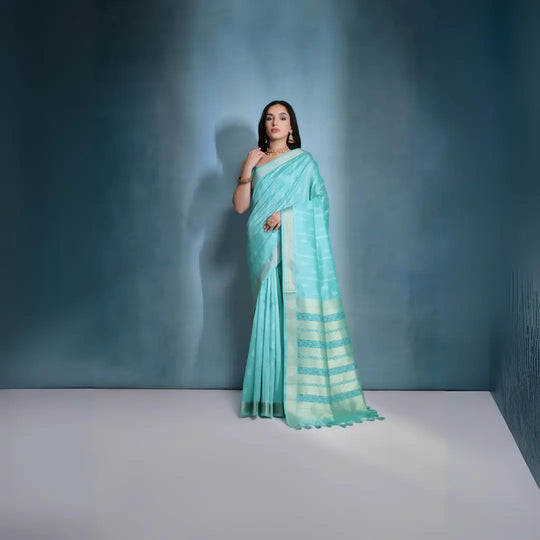 A woman models a serene Sky Blue Soft Raw Silk Saree. The saree body features subtle tone-on-tone Ikat weaving. The pallu is highlighted by wide horizontal bands of metallic silver/gold Zari weaving. She wears a blue sleeveless blouse and gold Kundan jewelry.
