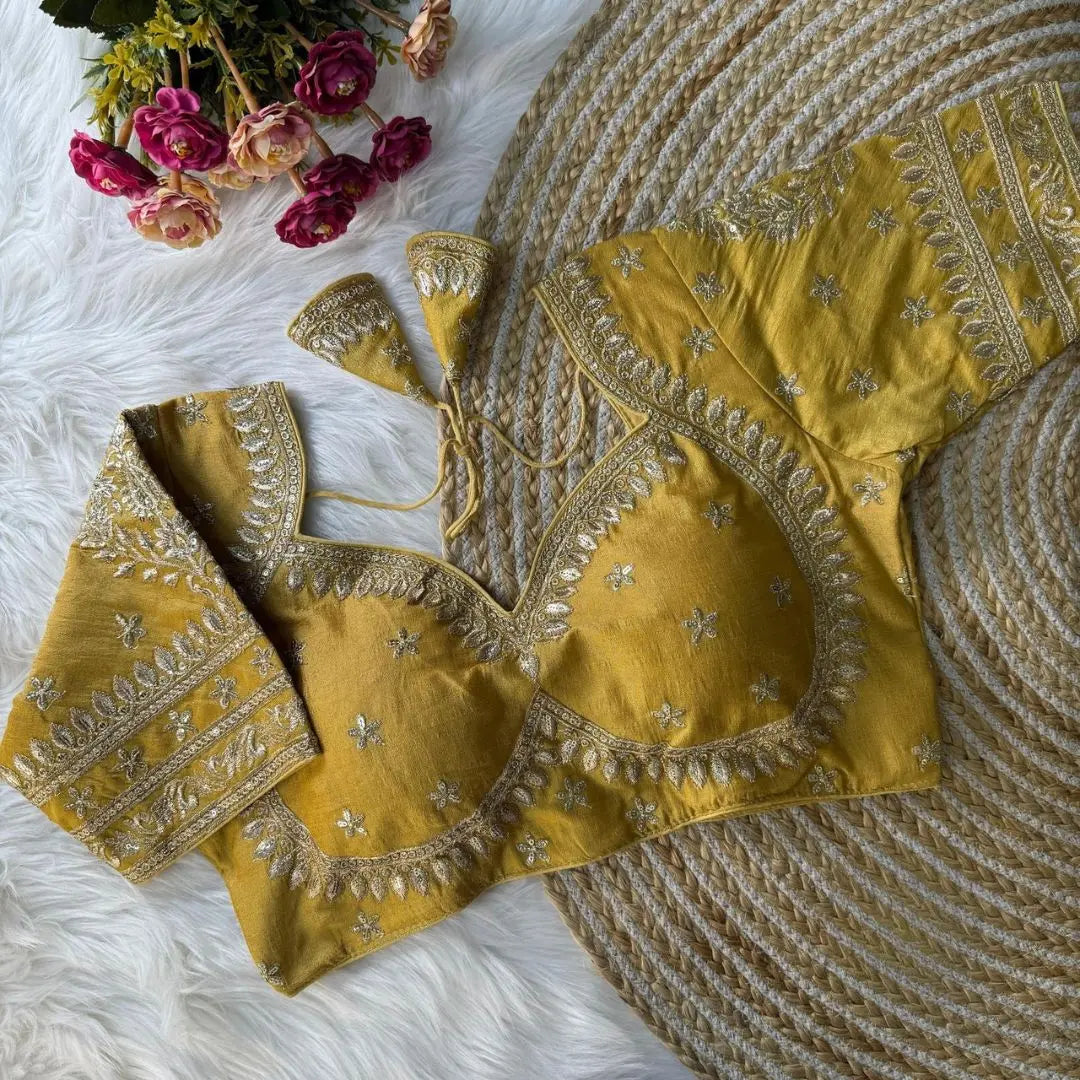 Mustard yellow embroidered silk blouse with intricate gold zari floral motifs and a tie-back design, displayed on a white fur background with a bunch of pink and peach flowers in the corner.