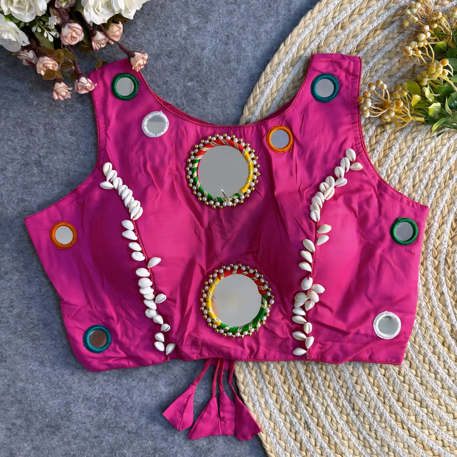 Bright fuchsia Indian blouse adorned with round mirrors and cowrie shell embellishments laid flat on a textured background