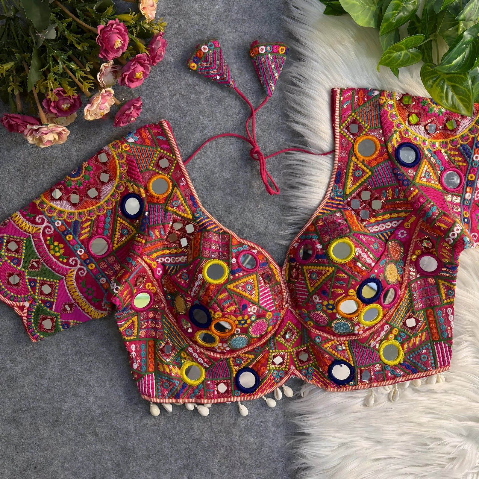 Traditional vibrant blouse featuring intricate multicolor mirror work and geometric embroidery in pink, yellow, green, orange, and blue, adorned with large and small round mirrors, beaded shell trim, and tasseled tie-back, displayed flat on a grey surface with artificial pink flowers and lush green foliage at the corners.