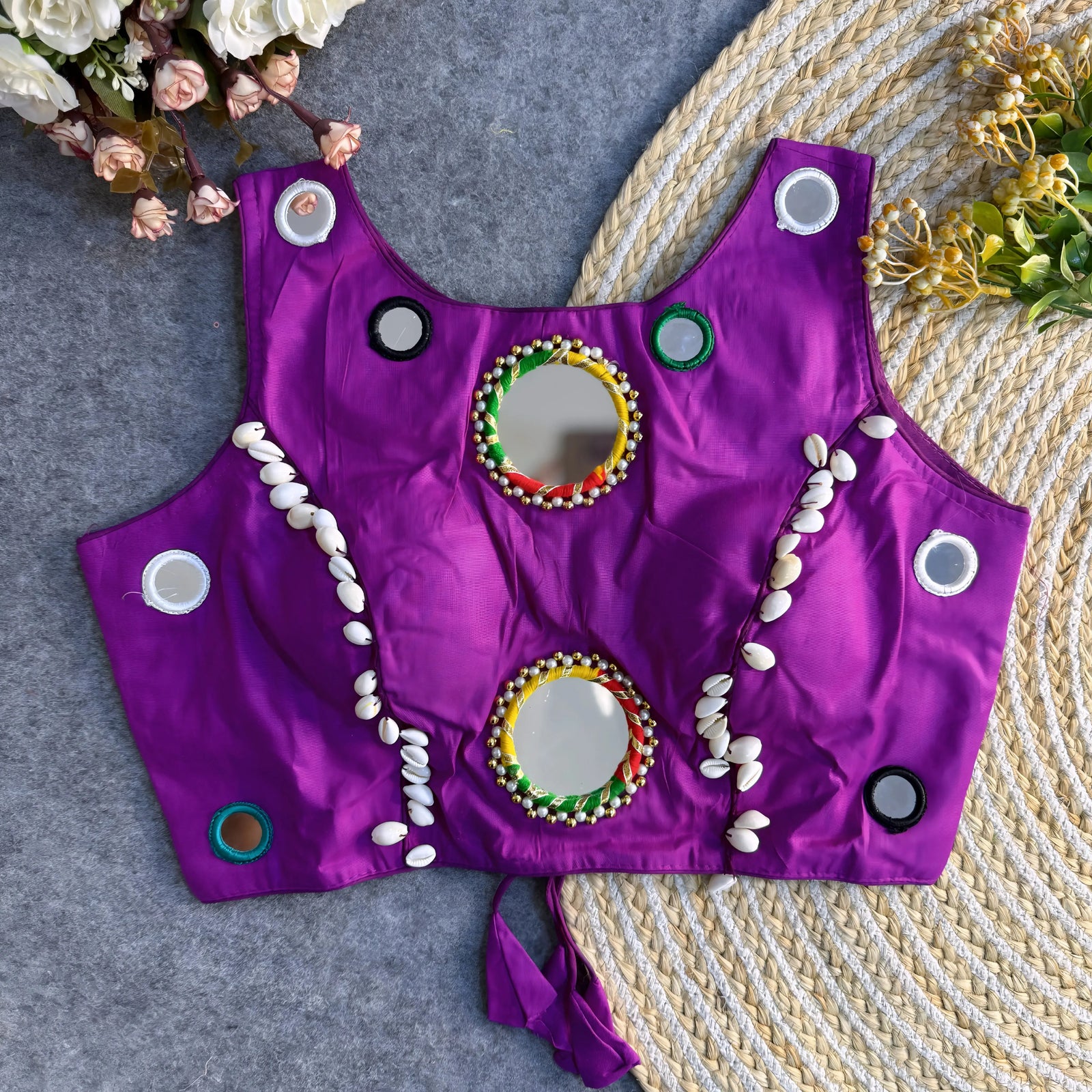 Bright purple ethnic blouse featuring circular mirror work, cowrie shell embellishments, and multicolor bead embroidery displayed on a woven mat.
