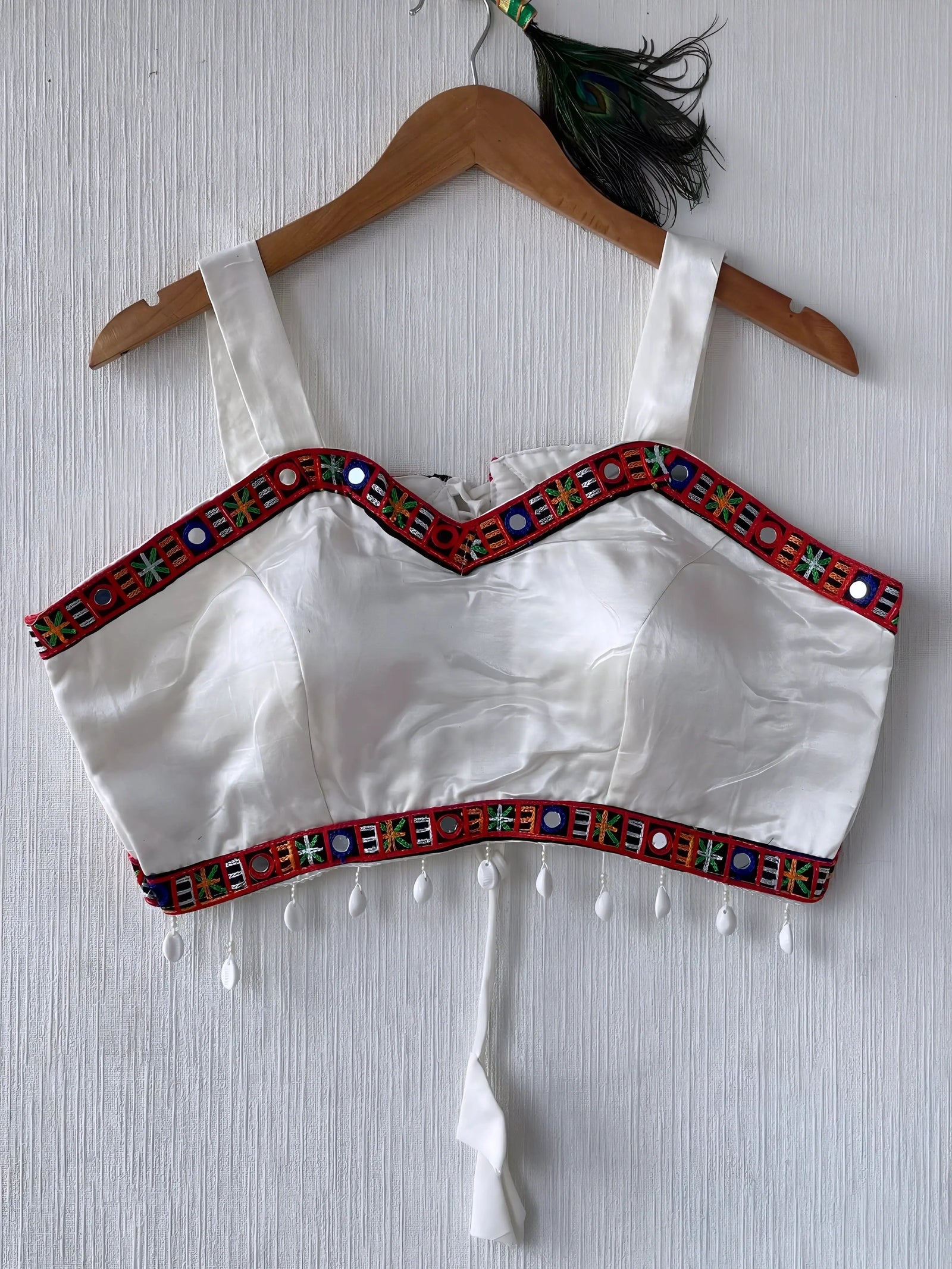 White ethnic blouse featuring vibrant mirror work and multicolored embroidered geometric border in red, green, and blue, adorned with white pearl drop tassels and a tie-up waist detail. Displayed on a wooden hanger against a textured off-white wall with peacock feathers for a festive and traditional look.
