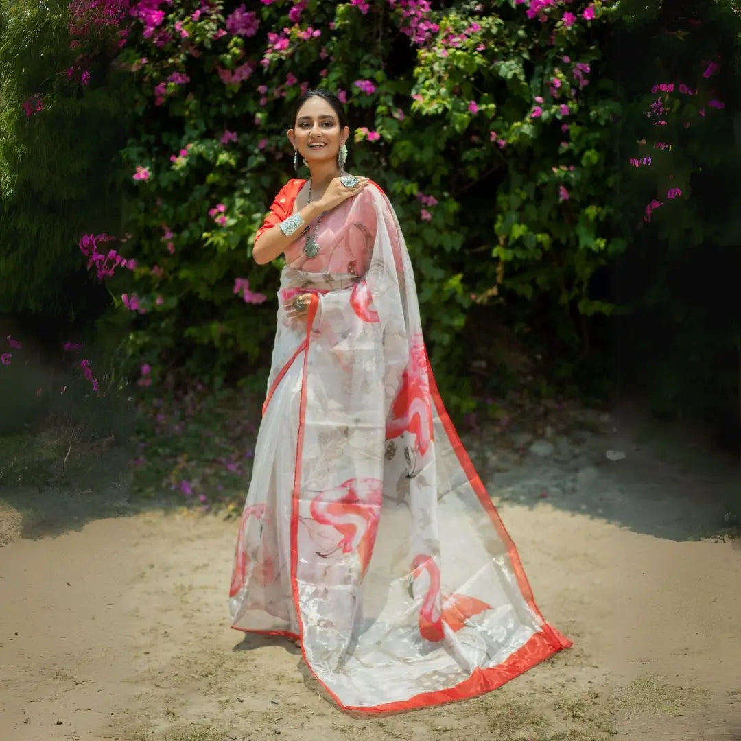 White transparent organza saree with coral flamingo pattern print orange red border paired with matching blouse turquoise silver jewelry outdoor bougainvillea background summer collection