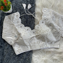 White bridal blouse with scallop sleeves, featuring circular mirror work and silver sequin stripes, decorative borders on sleeves and hem, and tie-up tassels, displayed on a textured grey and white fur background with a small potted plant nearby
