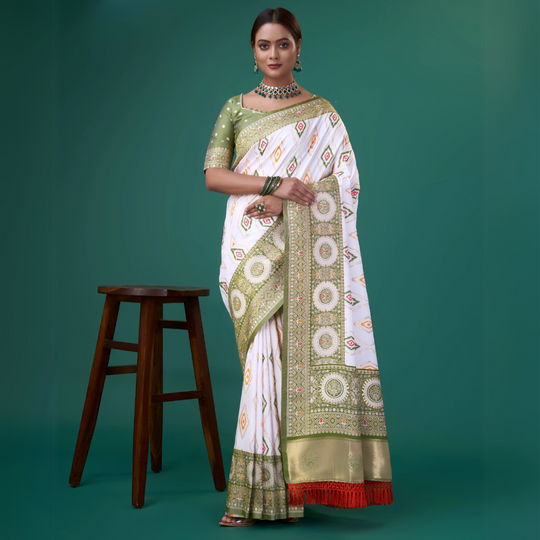 A woman models a striking White Patola Silk Saree featuring colorful Ikat-style motifs. The wide border and pallu are heavily woven in sage green Zari with intricate traditional medallion patterns. She wears a light green blouse and a heavy green gemstone choker necklace.