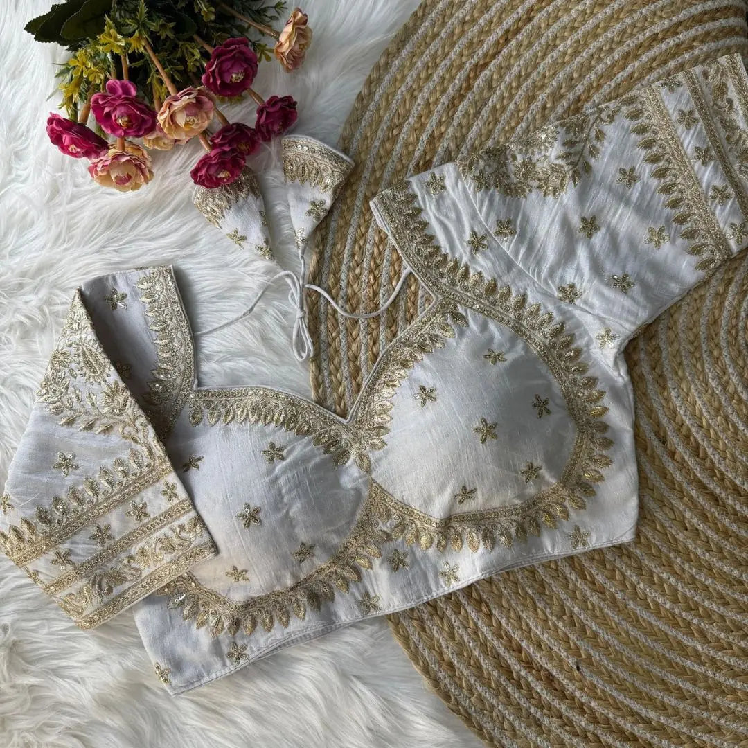 White blouse with intricate gold zari embroidery featuring floral and leaf motifs, decorative borders on sleeves and neckline, and tie-up tassels, displayed on a textured mat with pink flowers in the background