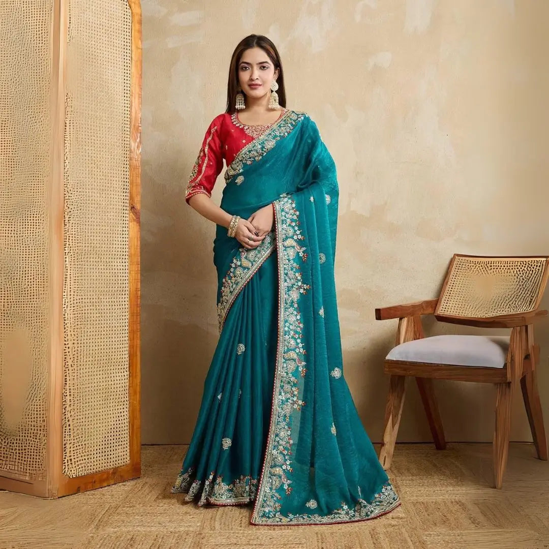 A woman models a vibrant Dark Pink Chiffon Silk Saree with scattered embellishments. The saree has a wide, intricate silver embroidery border. She wears a heavily embroidered dark green blouse and accessorizes with silver jewelry, posing indoors.
