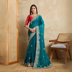 A woman models a vibrant Dark Pink Chiffon Silk Saree with scattered embellishments. The saree has a wide, intricate silver embroidery border. She wears a heavily embroidered dark green blouse and accessorizes with silver jewelry, posing indoors.