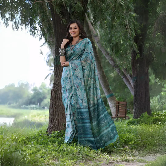 A woman models a Soft Tussar Silk Saree in a Rama Blue/Teal color, covered in an all-over artistic print. The pallu features dense Zari weaving in a striped, traditional pattern. She wears a teal blouse and a substantial set of oxidized silver jewelry, posing against a large tree.