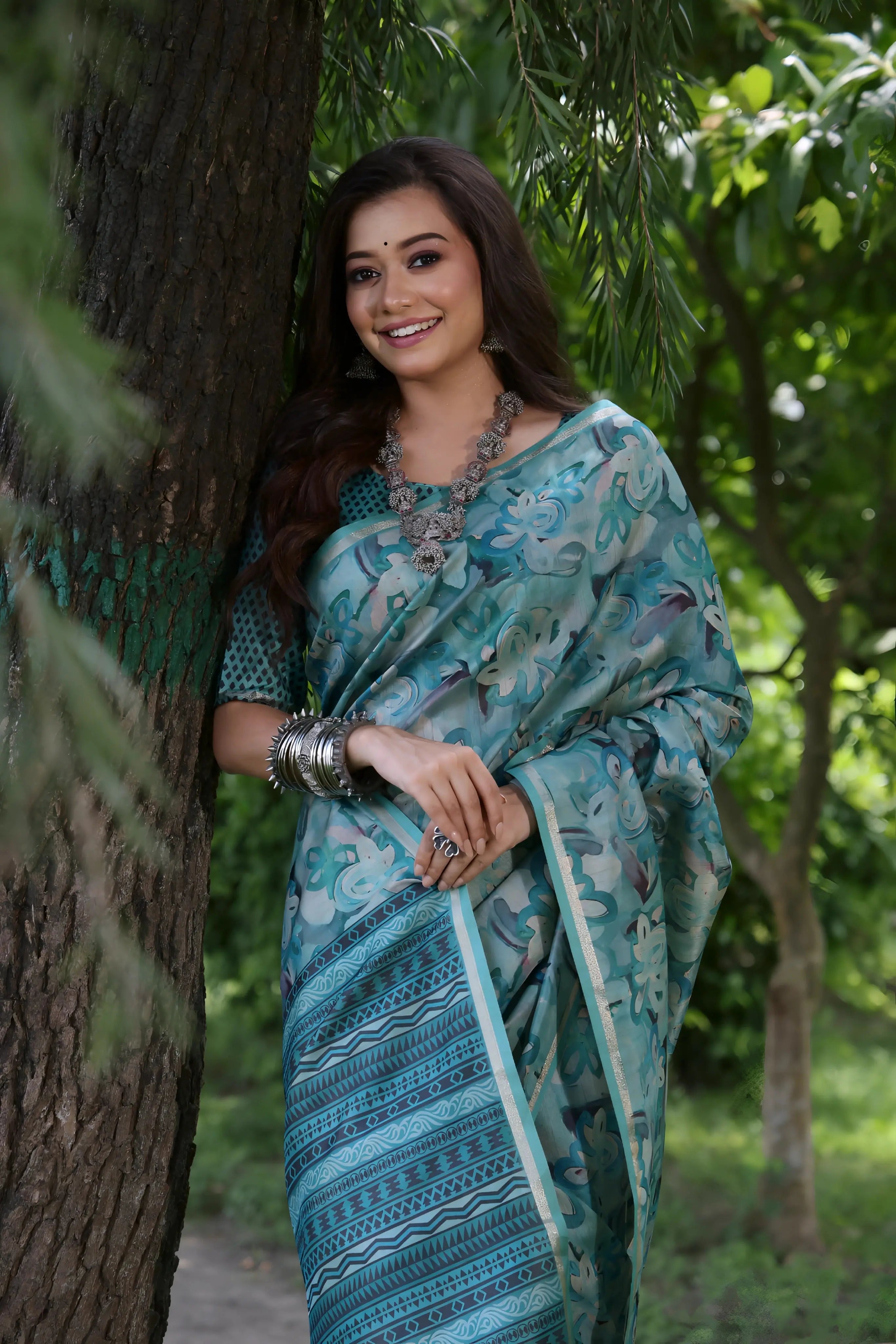 A woman models a Soft Tussar Silk Saree in a Rama Blue/Teal color, covered in an all-over artistic print. The pallu features dense Zari weaving in a striped, traditional pattern. She wears a teal blouse and a substantial set of oxidized silver jewelry, posing against a large tree.