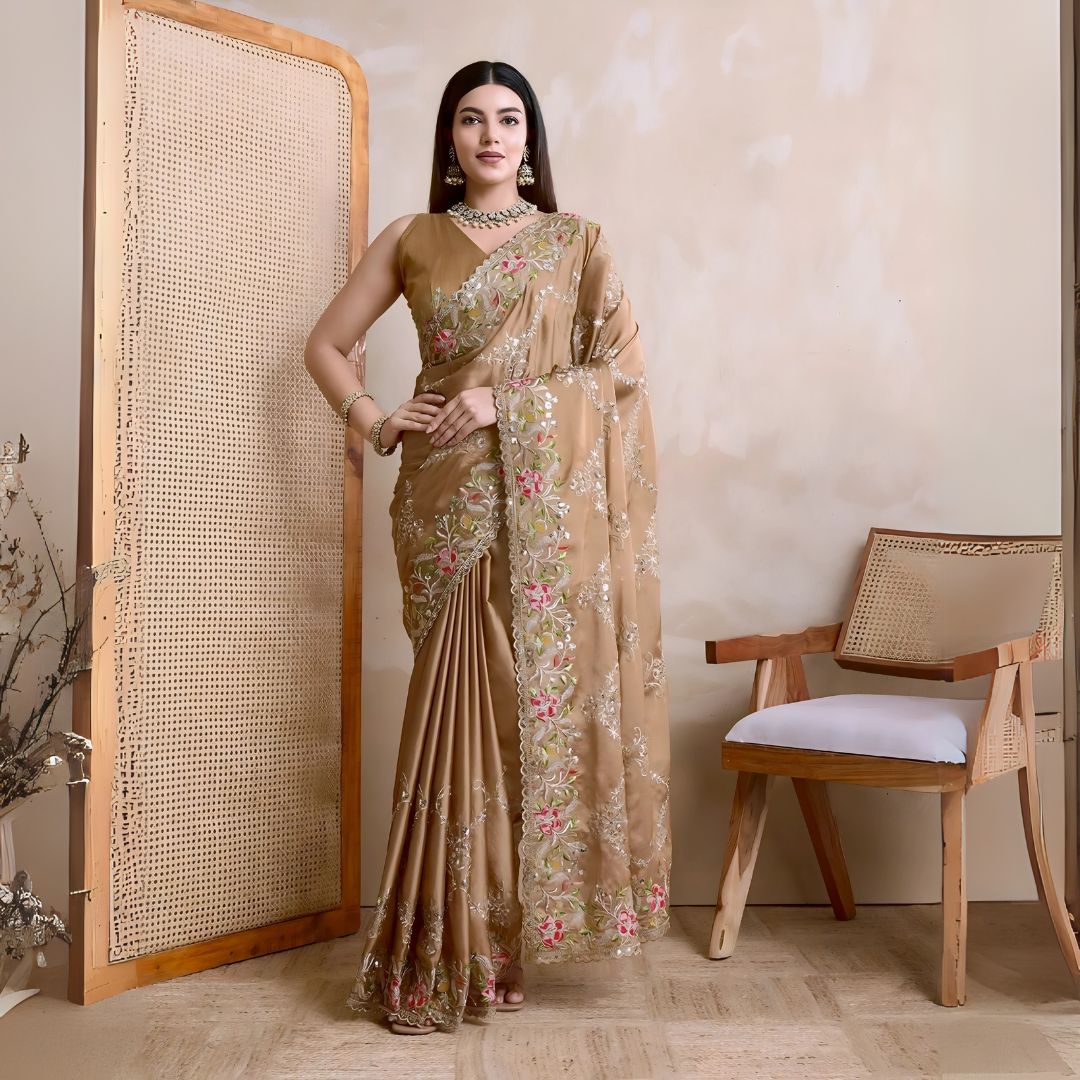 A woman models an opulent Beige Rangoli Silk Saree with scattered sequins. The saree is bordered by wide, dense floral embroidery in pink, red, and green threadwork. She wears a beige sleeveless blouse and a heavy green gemstone necklace.