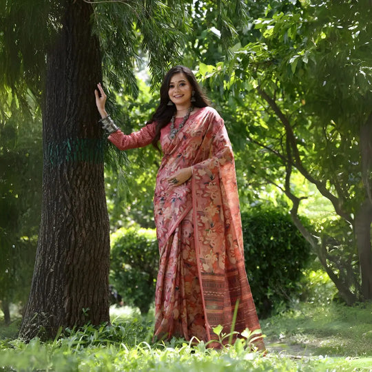 A woman models a Soft Tussar Silk Saree in a dusty maroon color, covered in an elaborate all-over artistic print. The saree has a distinct woven Zari border. She wears a matching long-sleeve blouse and an oxidized silver necklace, posing outdoors in a garden.