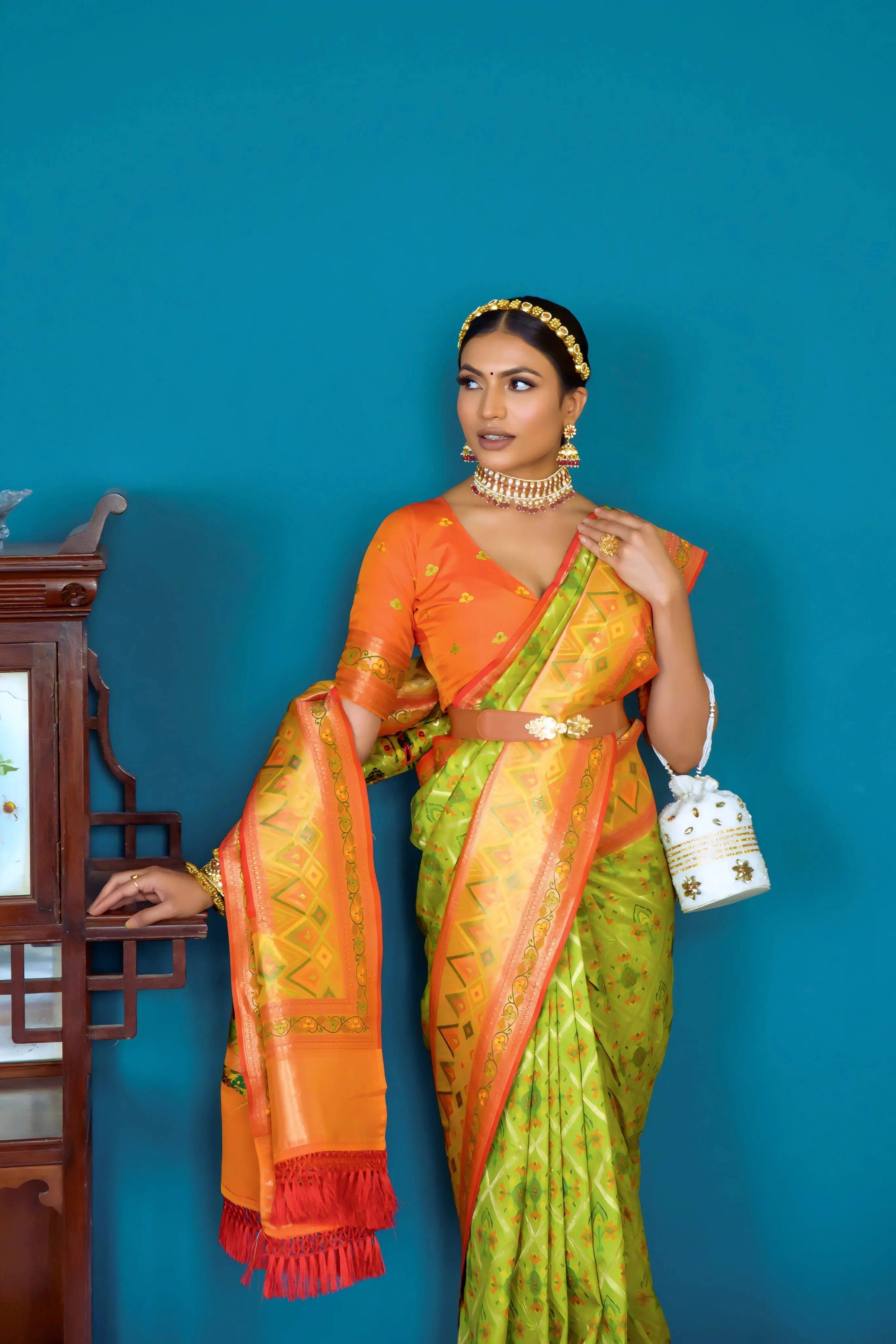 A woman models a striking Lime Green Banarasi Silk Saree with all-over Zari weaving. The wide pallu and border contrast dramatically in Bright Orange with large traditional Zari weaving patterns. She wears an orange blouse, traditional gold jewelry, and a metallic waist belt, posing indoors.