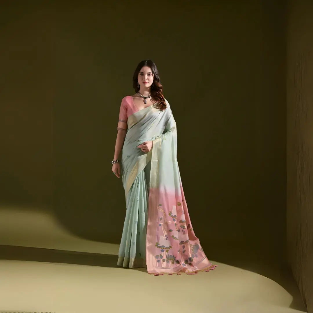 A woman models a Soft Muga Cotton Saree. The saree's body is pale sea green with a gold Zari border. The pallu dramatically transitions to a soft rose pink color featuring woven motifs. She wears a pink blouse and a dark gemstone choker necklace.
