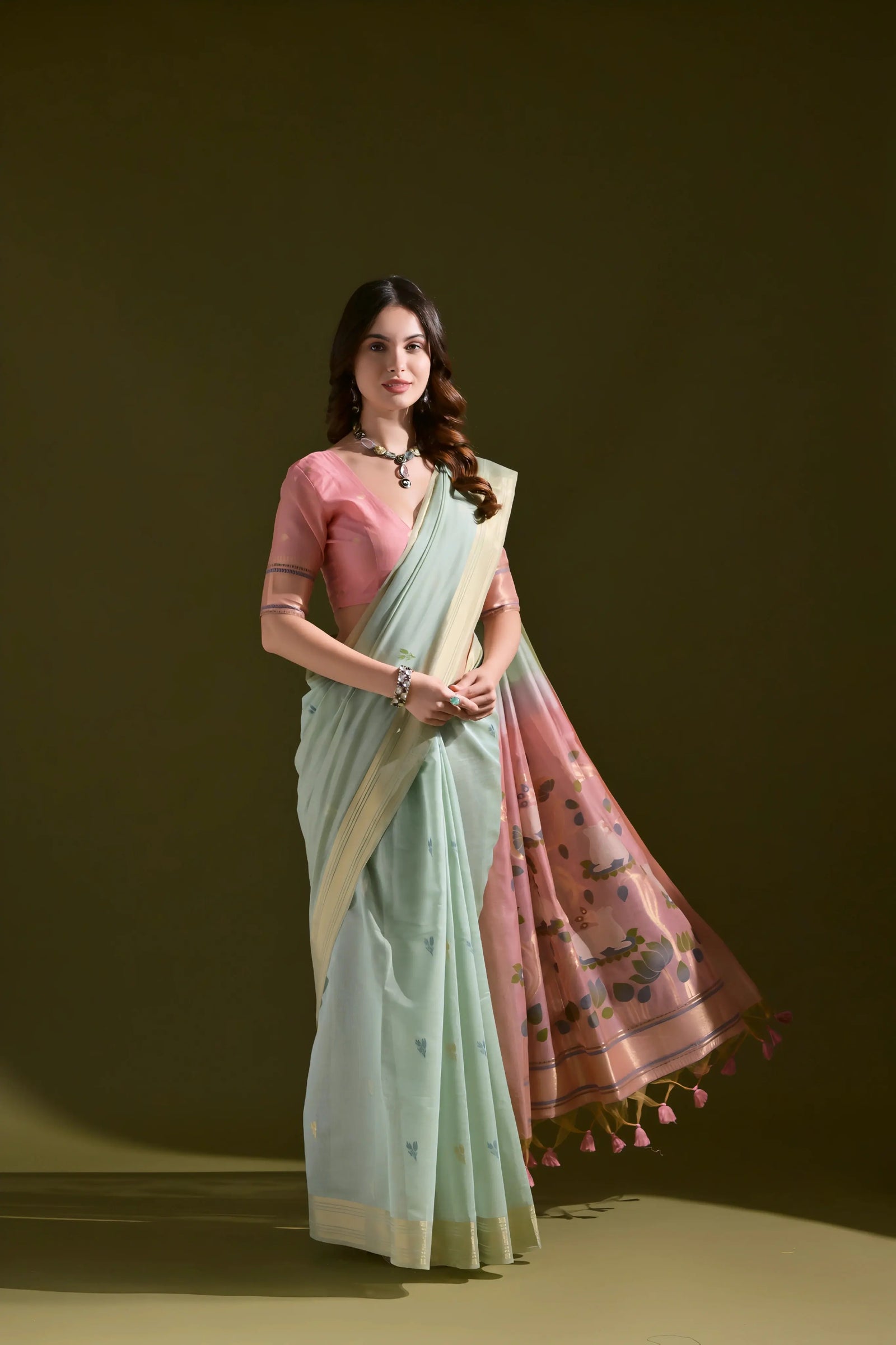 A woman models a Soft Muga Cotton Saree. The saree's body is pale sea green with a gold Zari border. The pallu dramatically transitions to a soft rose pink color featuring woven motifs. She wears a pink blouse and a dark gemstone choker necklace.