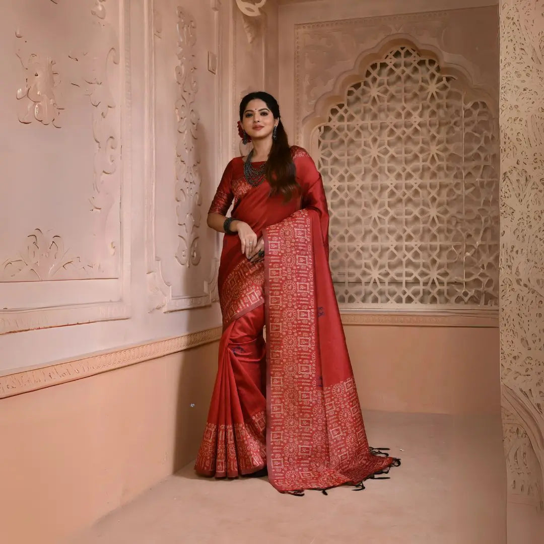Woman wearing a maroon silk saree with intricate geometric border and pallu, paired with traditional jewelry, standing in a heritage-inspired room with ornate wall carvings and a decorative lattice window.