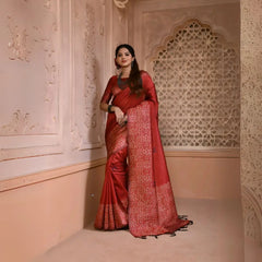 Woman wearing a maroon silk saree with intricate geometric border and pallu, paired with traditional jewelry, standing in a heritage-inspired room with ornate wall carvings and a decorative lattice window.