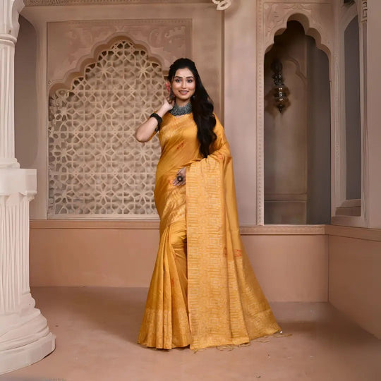 A woman models a vibrant Yellow Banglori Raw Silk Saree. The pallu and border feature dense, tone-on-tone Kalamkari-style weaving in gold Zari. She wears a yellow blouse and heavy dark gemstone jewelry, posing in an ornate, traditional setting.