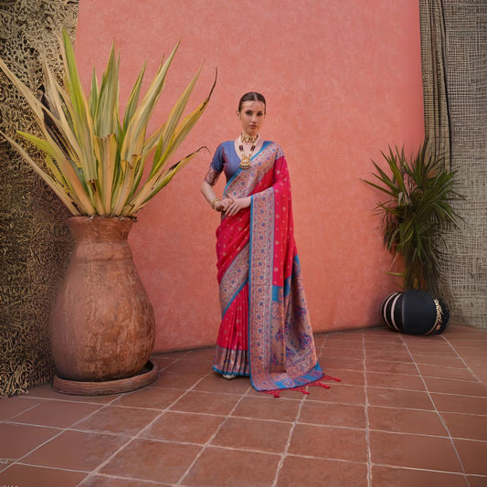 A woman models a striking Soft Banarasi Silk Saree. The saree is deep pink with gold Zari weaving across the body, featuring an ornate border and pallu with contrasting blue accents. She pairs it with a short-sleeve blue blouse and heavy traditional gold jewelry.