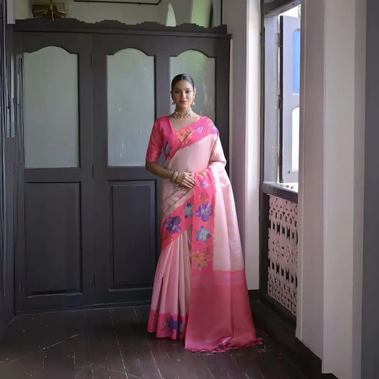 A woman models a graceful Soft Silk Saree. The saree body is pale pink with a wide hot pink border. The shoulder and pallu area feature large, vibrant printed floral motifs in blue and purple. She pairs it with a pink blouse and a gold choker necklace.