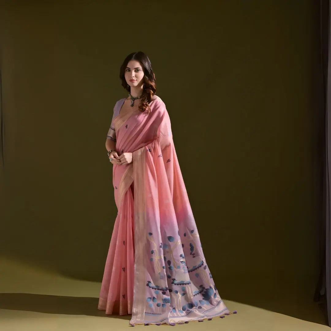 A woman models a Soft Muga Cotton Saree. The saree is soft pink with a gold Zari border. The pallu transitions into a pink and purple color scheme with large, woven blue and purple motifs. She pairs it with a light purple blouse and a choker necklace.