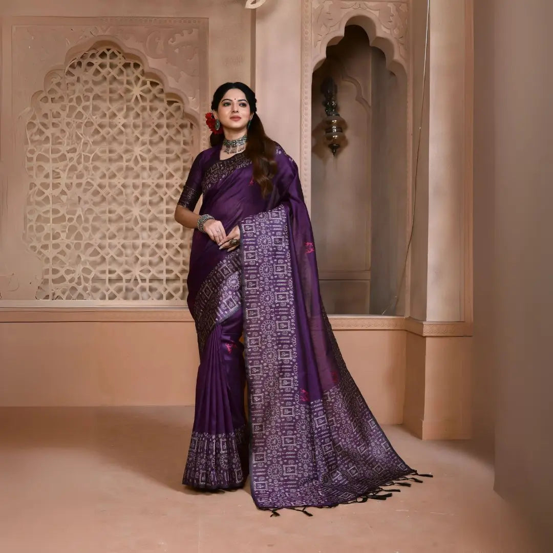 A woman models a striking Royal Purple Banglori Raw Silk Saree. The pallu and border feature dense, traditional Kalamkari-style weaving in metallic Zari thread. She wears a purple blouse and heavy green beaded jewelry, posing in an ornate, traditional setting.