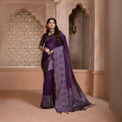 A woman models a striking Royal Purple Banglori Raw Silk Saree. The pallu and border feature dense, traditional Kalamkari-style weaving in metallic Zari thread. She wears a purple blouse and heavy green beaded jewelry, posing in an ornate, traditional setting.