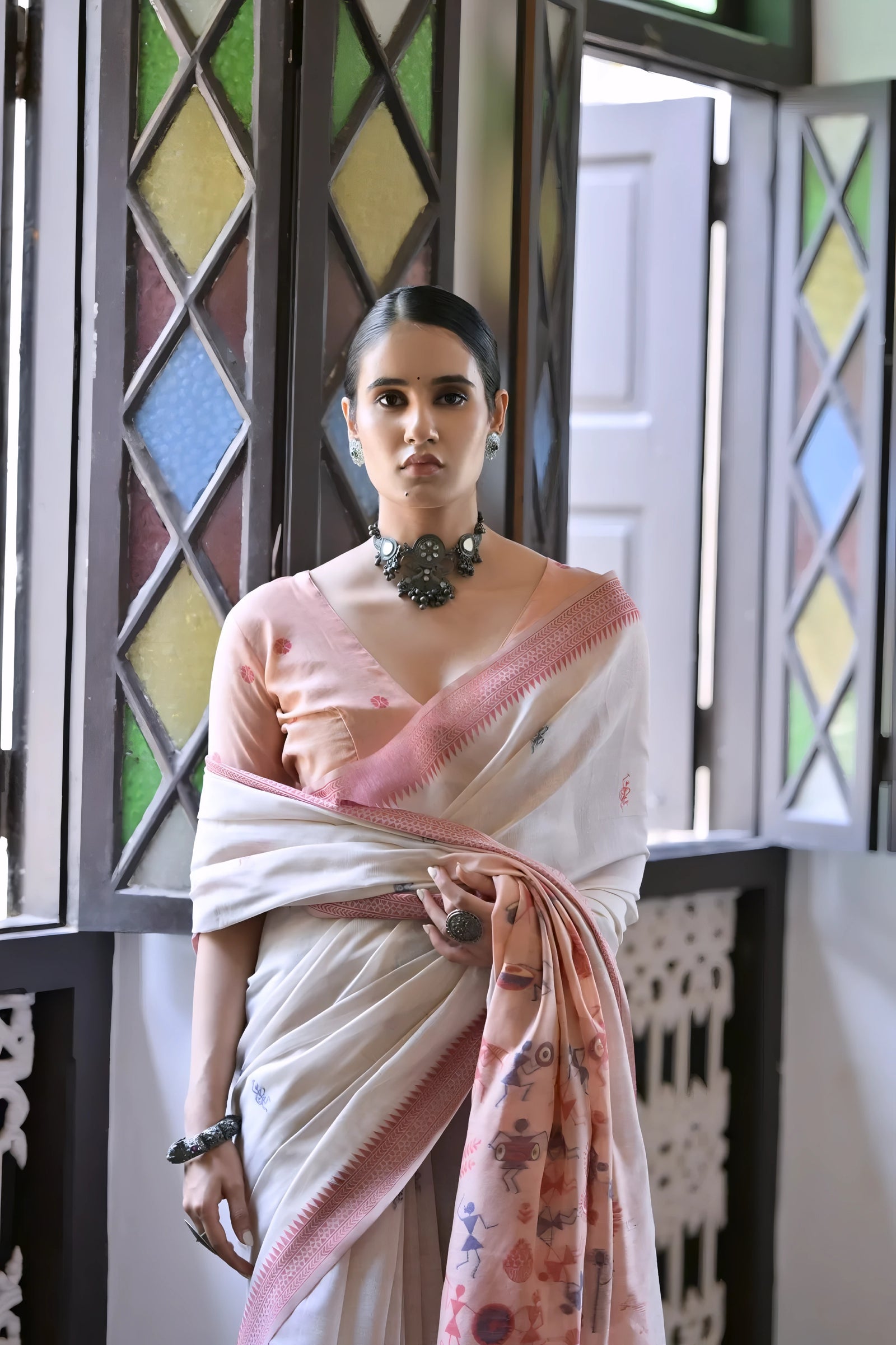 A woman models a graceful Off White Soft Muga Cotton Silk Saree. The saree has a dark pink woven border, and the pallu features a wide sky blue panel decorated with traditional Warli-style figurative motifs. She wears a light blue blouse and a dark oxidized choker necklace, posing indoors.